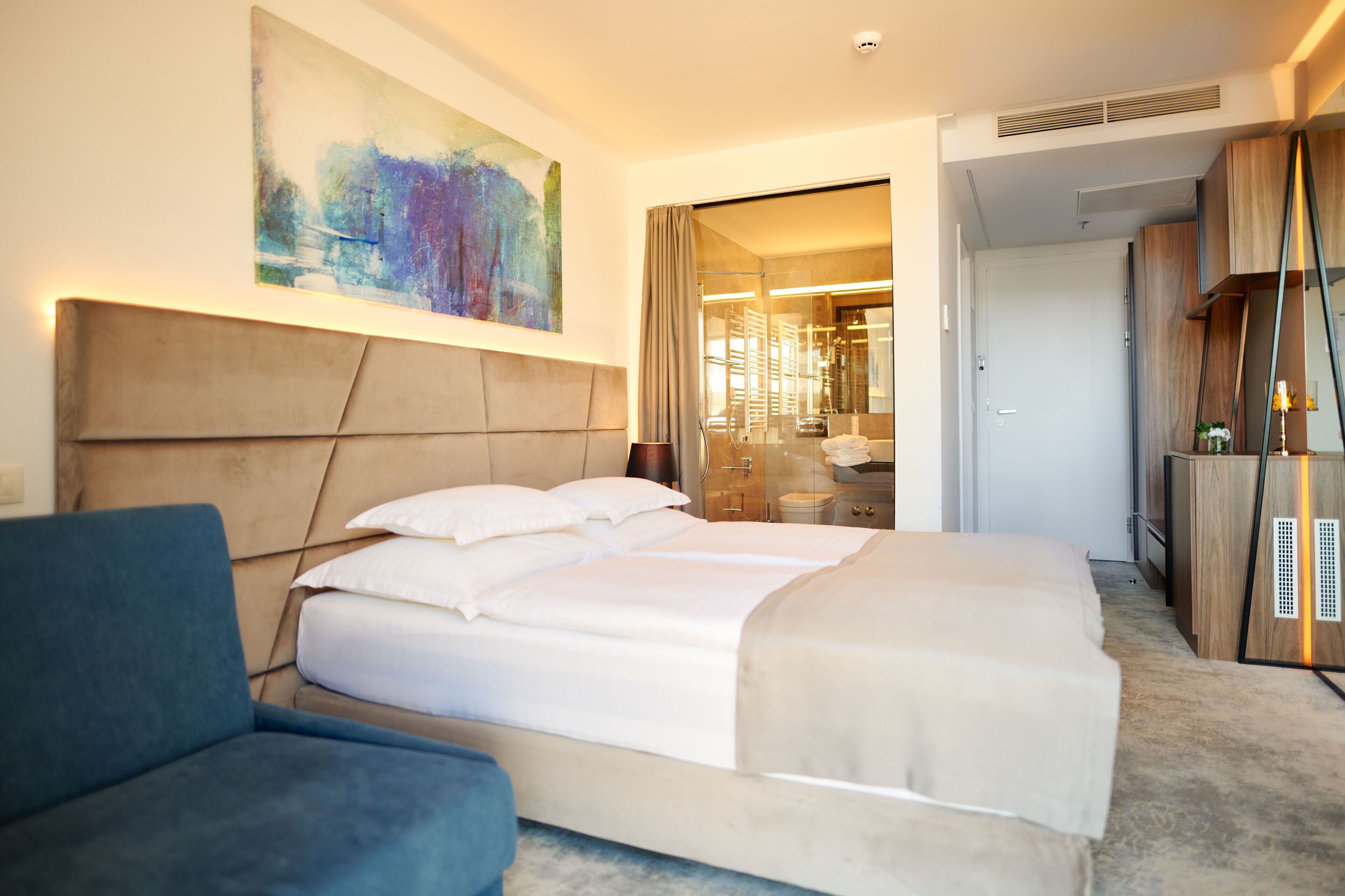 Une chambre d’hôtel moderne avec un grand lit, un fauteuil bleu et un grand tableau abstrait accroché au mur.
