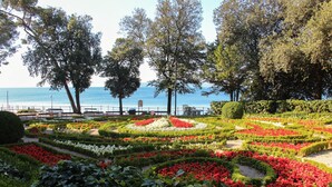 Un jardín ajardinado con diferentes parterres de flores y árboles, un sendero y vistas al mar.