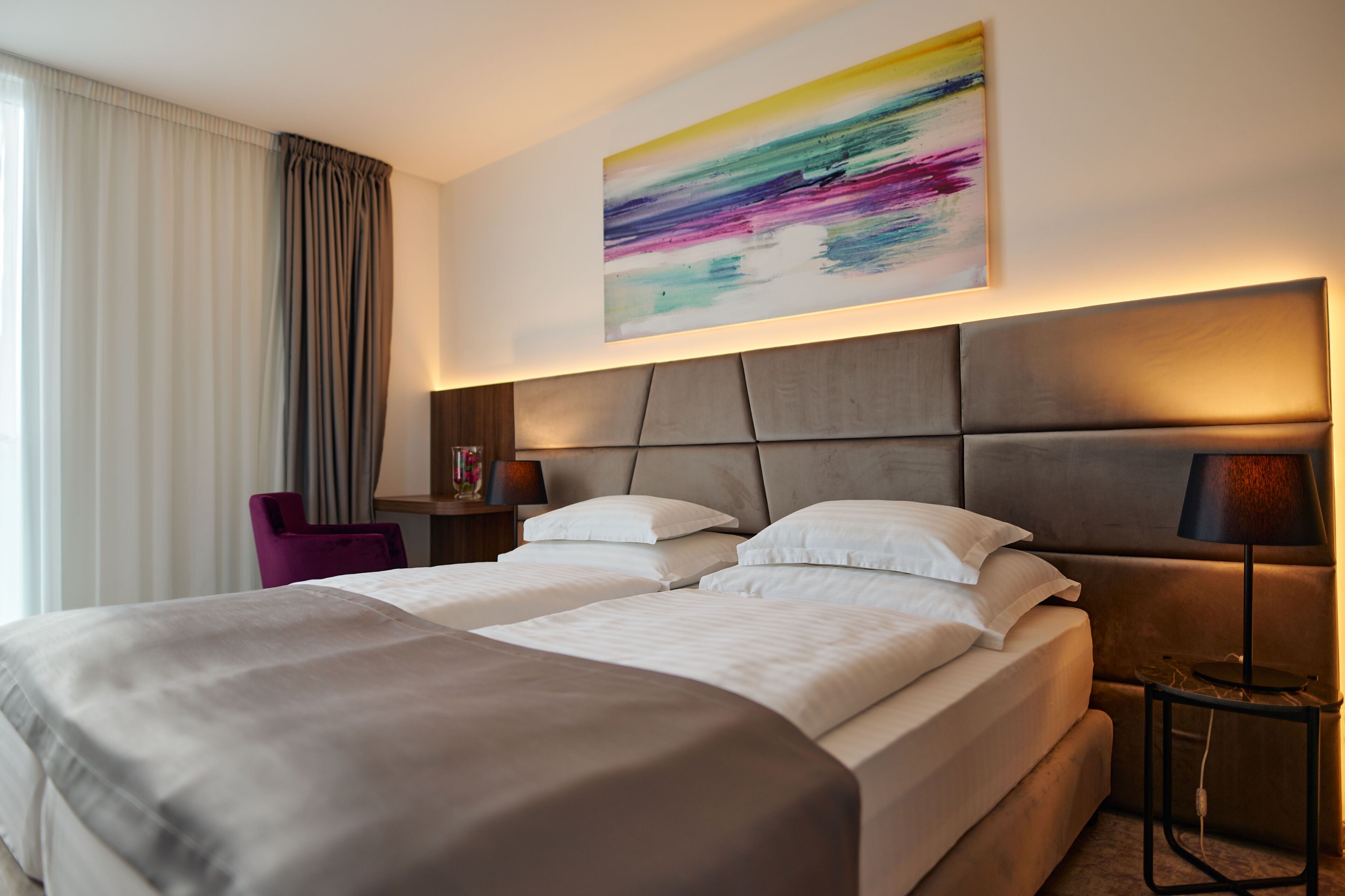 A hotel room with a large bed, a bedside table with a lamp, a chair, and a colorful abstract painting on the wall.