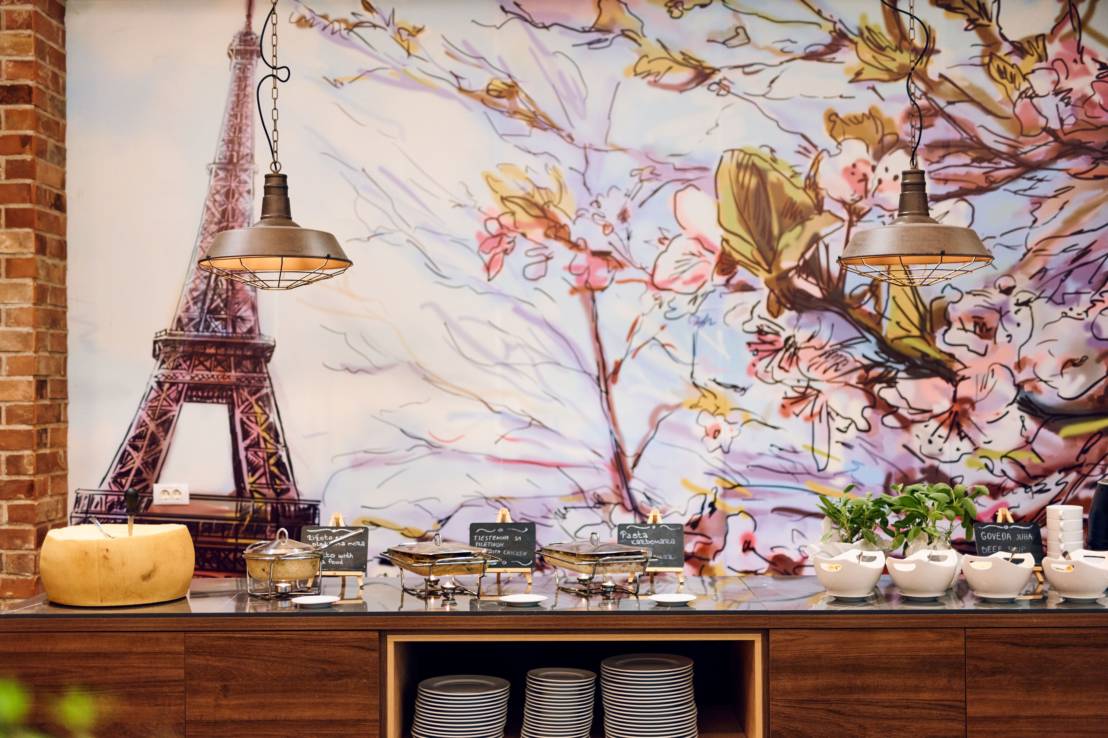 A buffet setup with a decorative Eiffel Tower backdrop, a hanging light fixture, and labeled serving dishes.