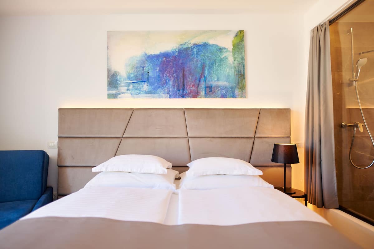A hotel room with a large bed, a headboard, a painting on the wall, a shower area, and a blue chair.