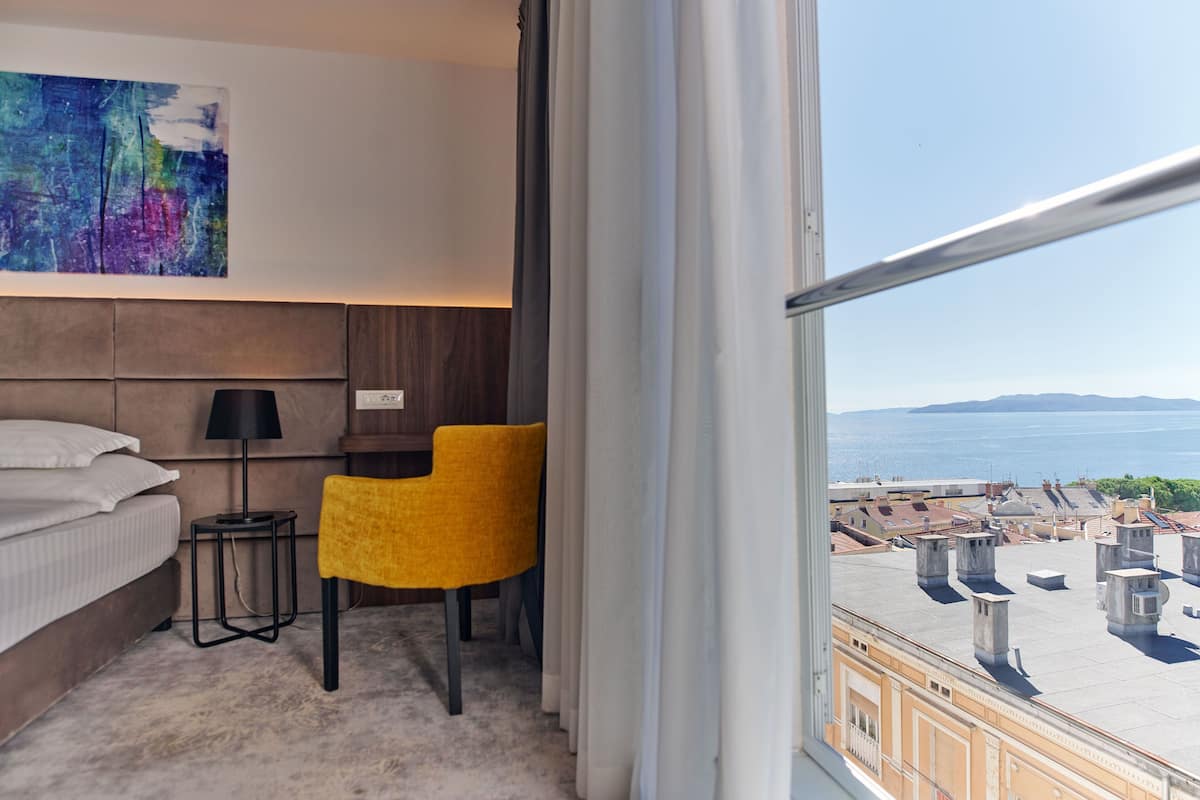 A hotel room with a bed, a desk, a chair, a lamp, and a large window with a view of the city and the sea.