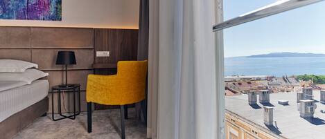 A hotel room with a bed, a desk, a chair, a lamp, and a large window with a view of the city and the sea.