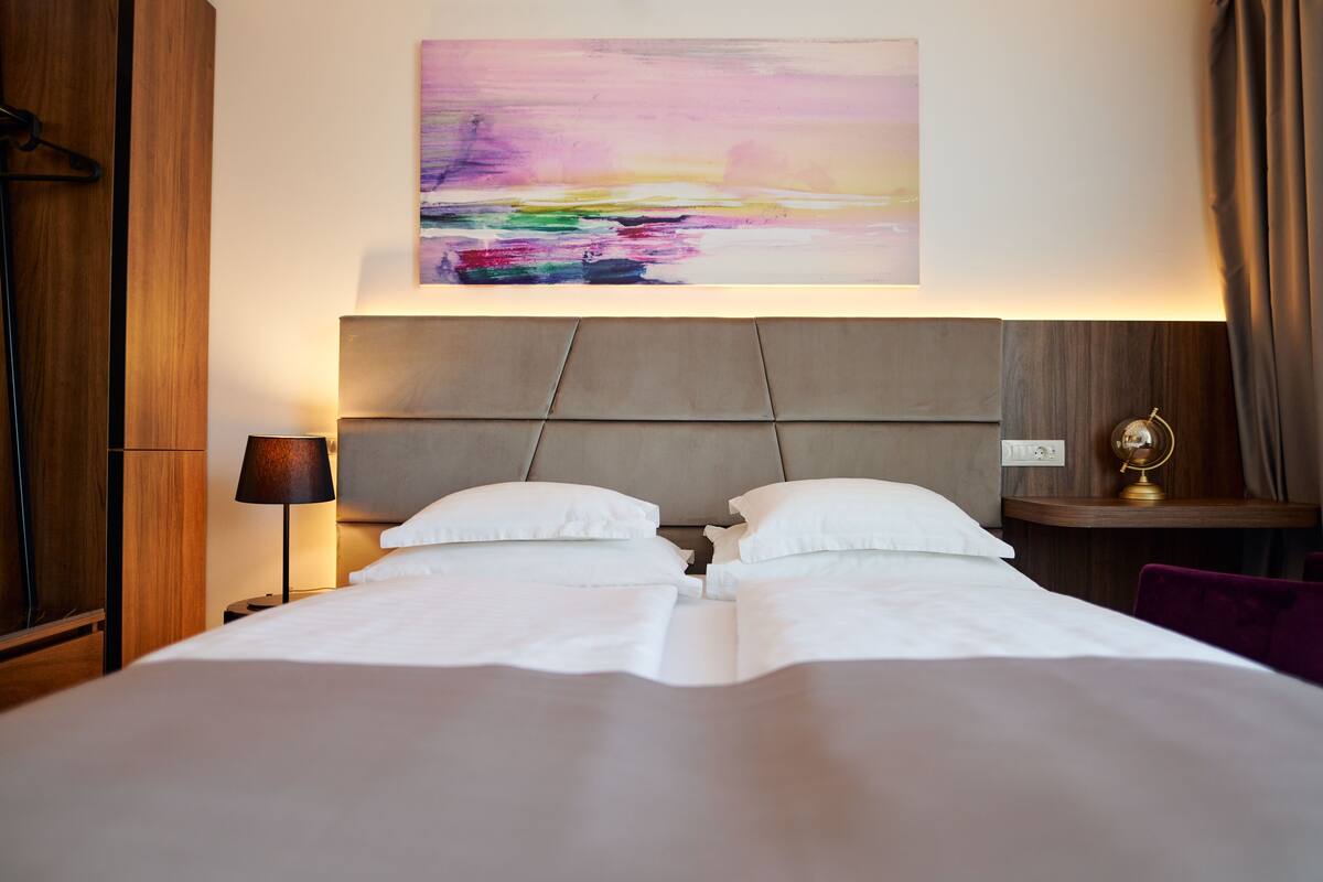 A hotel room with a large bed, a headboard, a bedside table with a lamp, and a wall-mounted abstract painting.