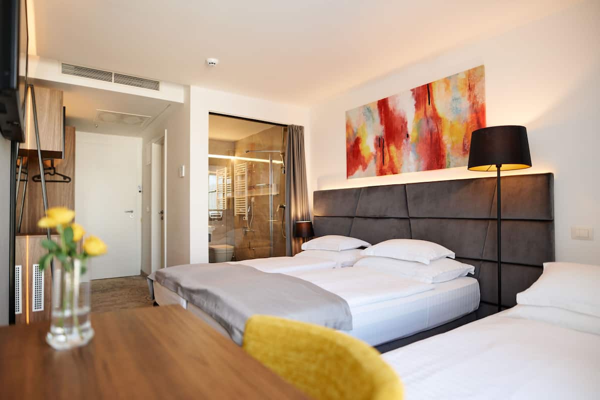 A hotel room with a bed, a bedside table with a vase of flowers, a lamp, and a colorful abstract painting on the wall.