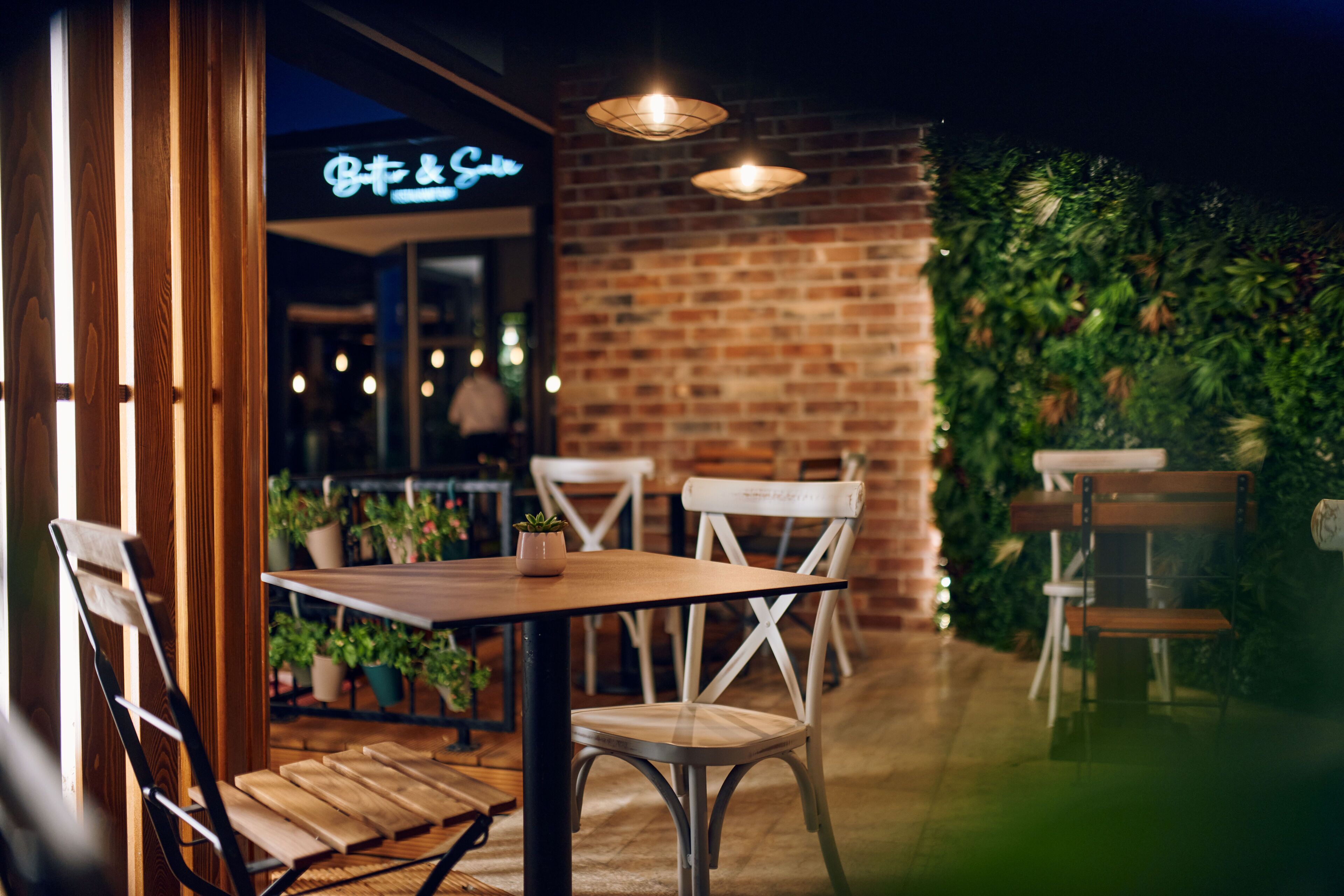 An outdoor seating area with wooden tables and chairs, potted plants, and a brick wall.
