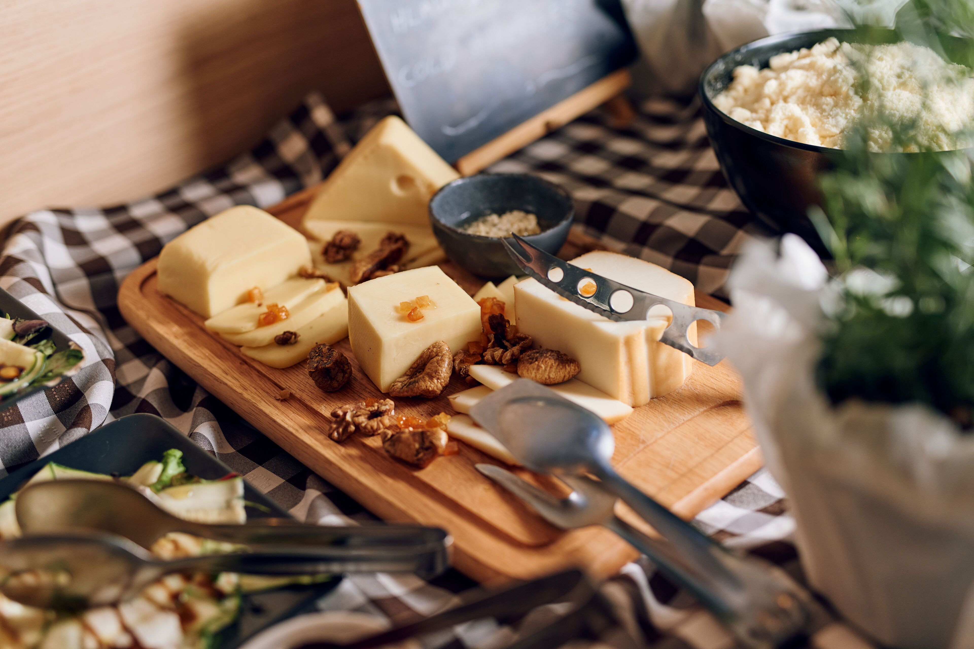 A wooden board with various cheeses and nuts, a bowl of salt, and a spoon on a checkered cloth.