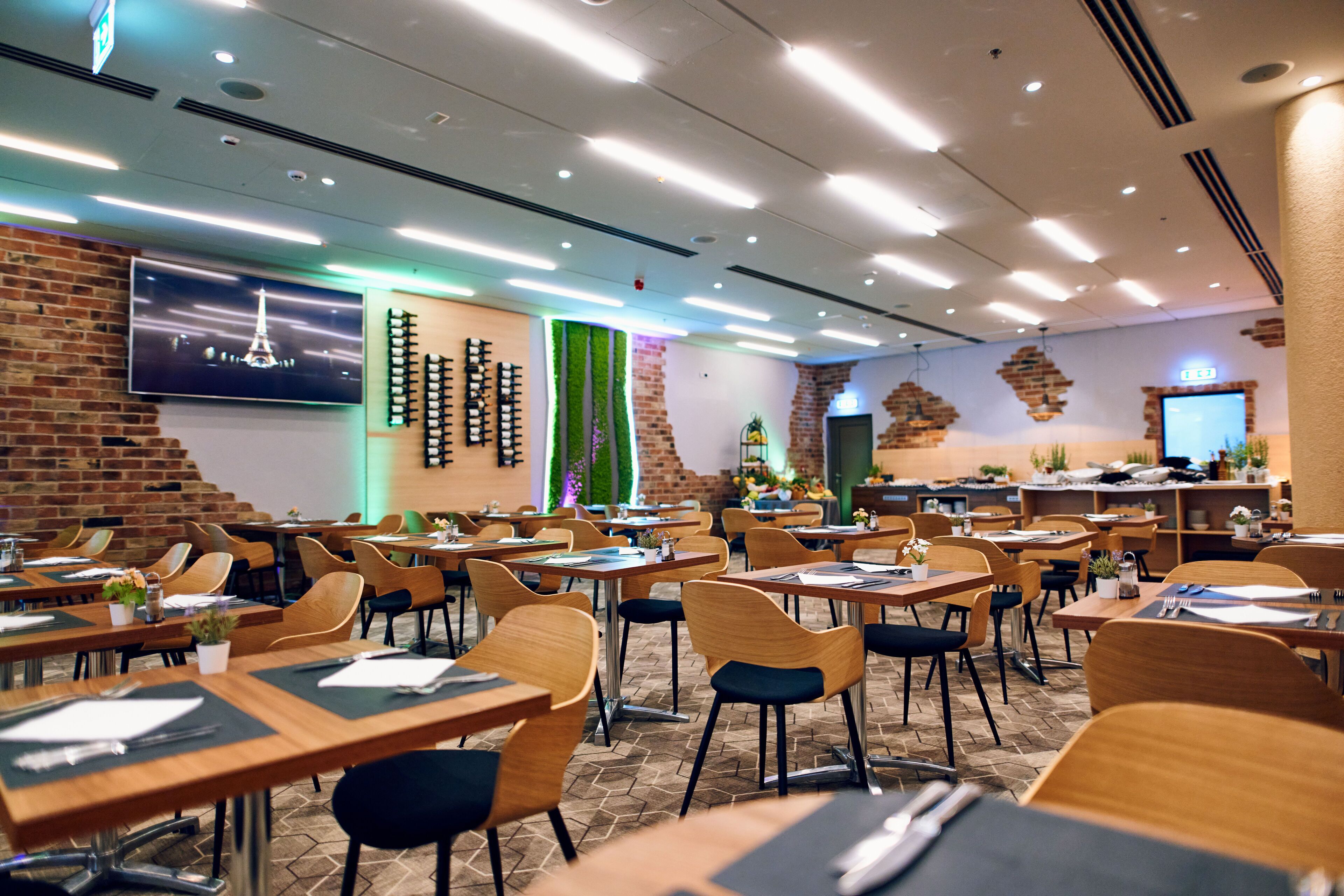 A restaurant with multiple tables and chairs, a brick wall, a TV screen, and a green wall with vertical plants.