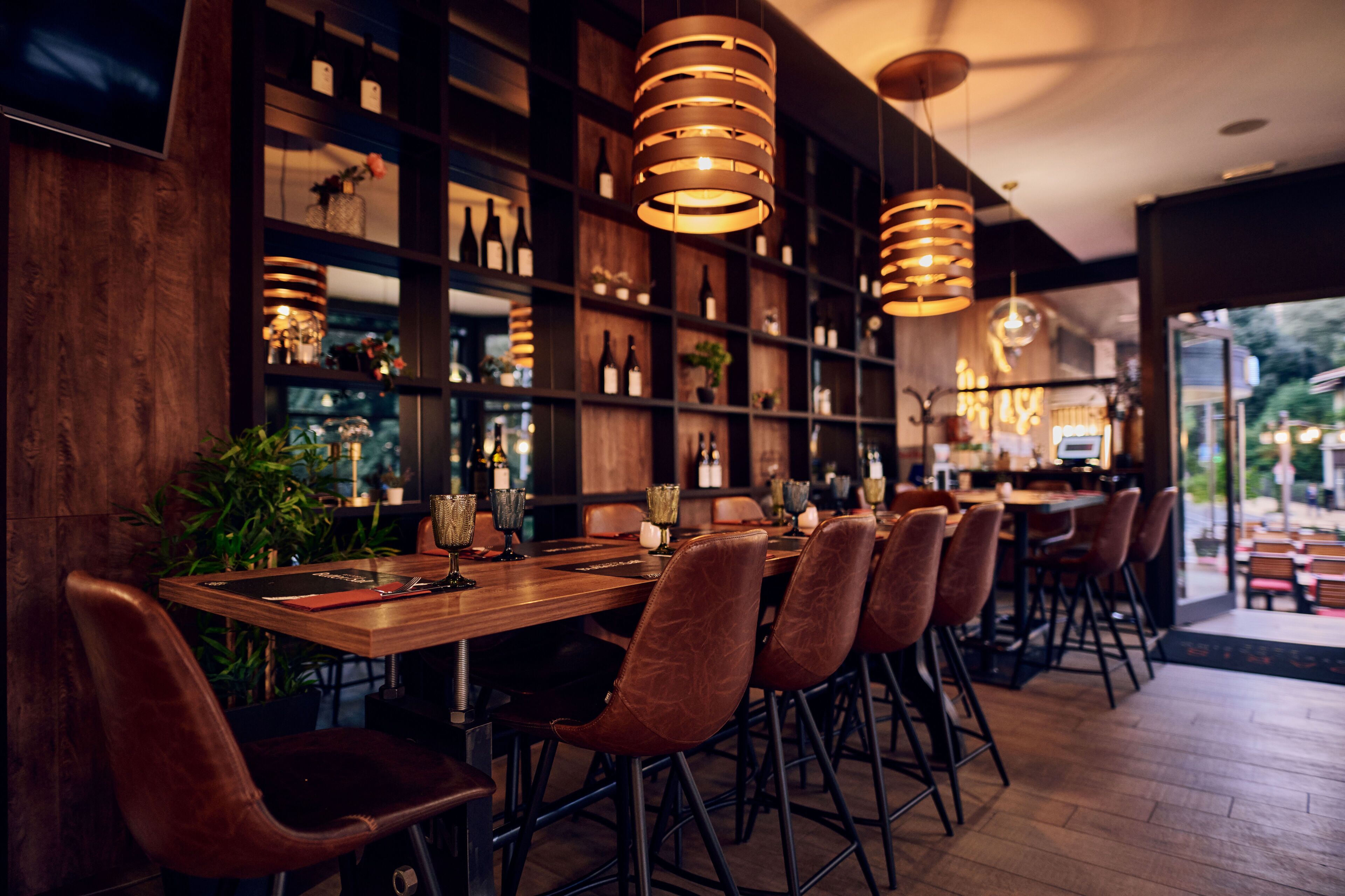 Un interior de restaurante con mesas de madera, sillas de cuero, lámparas colgantes y estantes llenos de botellas y copas.