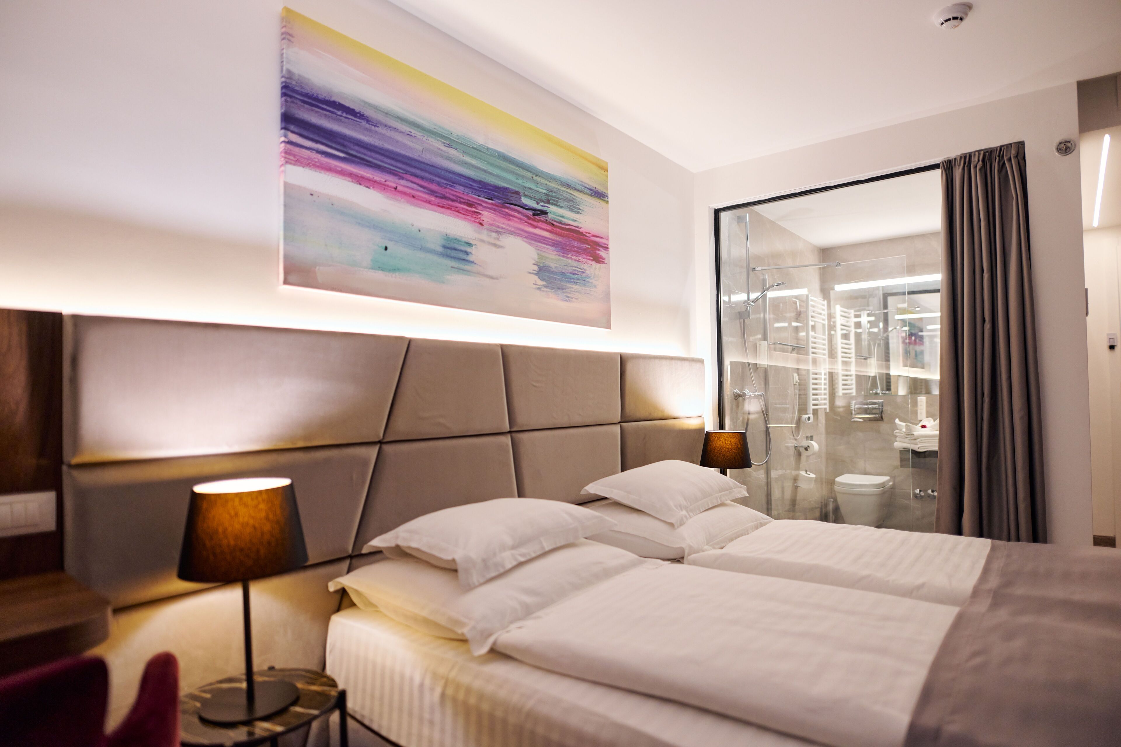 Ein modernes Hotelzimmer mit einem großen Bett, einem Kopfteil mit Lampenleuchte, einem Nachttisch mit Lampe und einer Duschkabine aus Glas.
