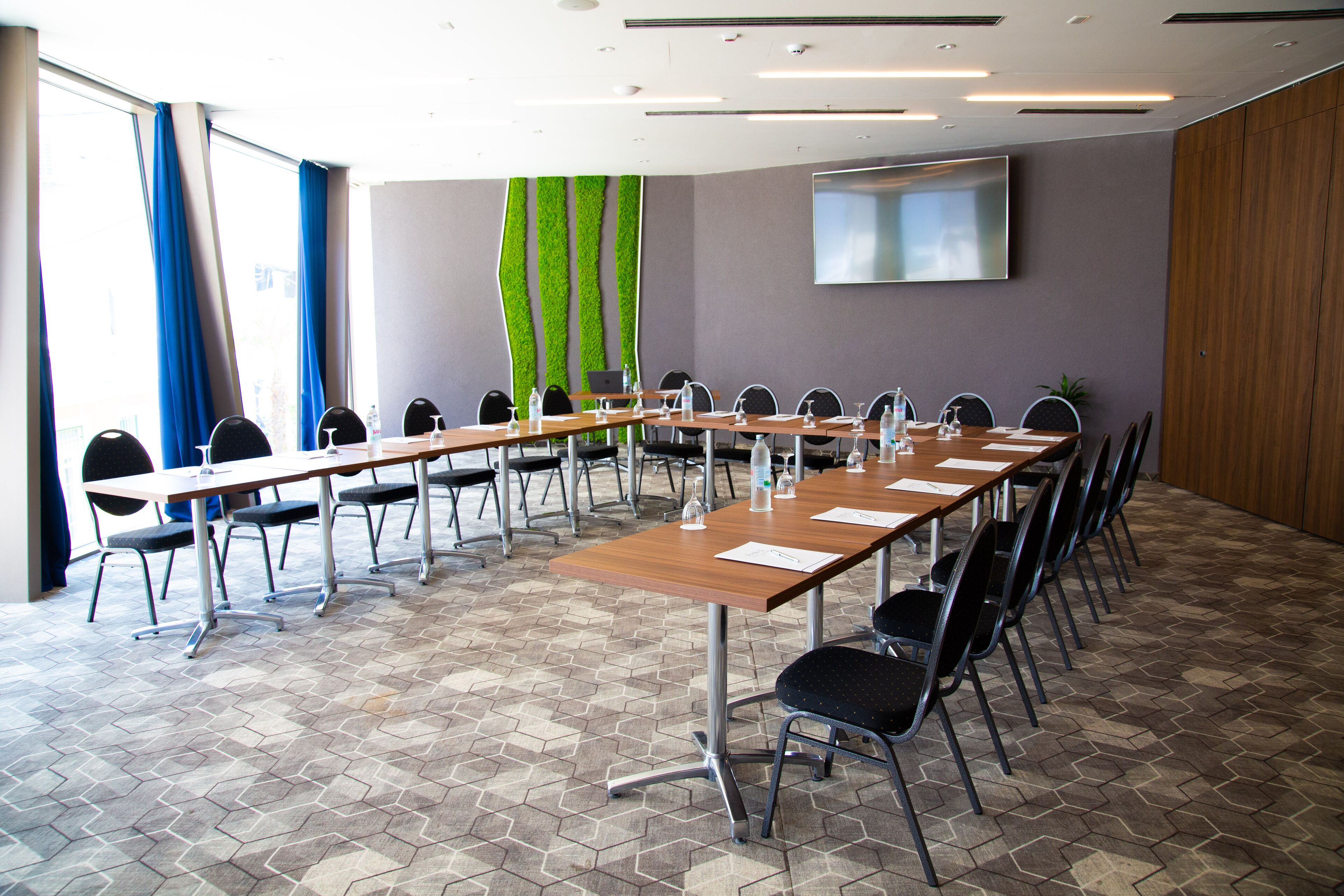 A conference room with long tables and chairs, a flat-screen TV on the wall, and a green vertical garden.