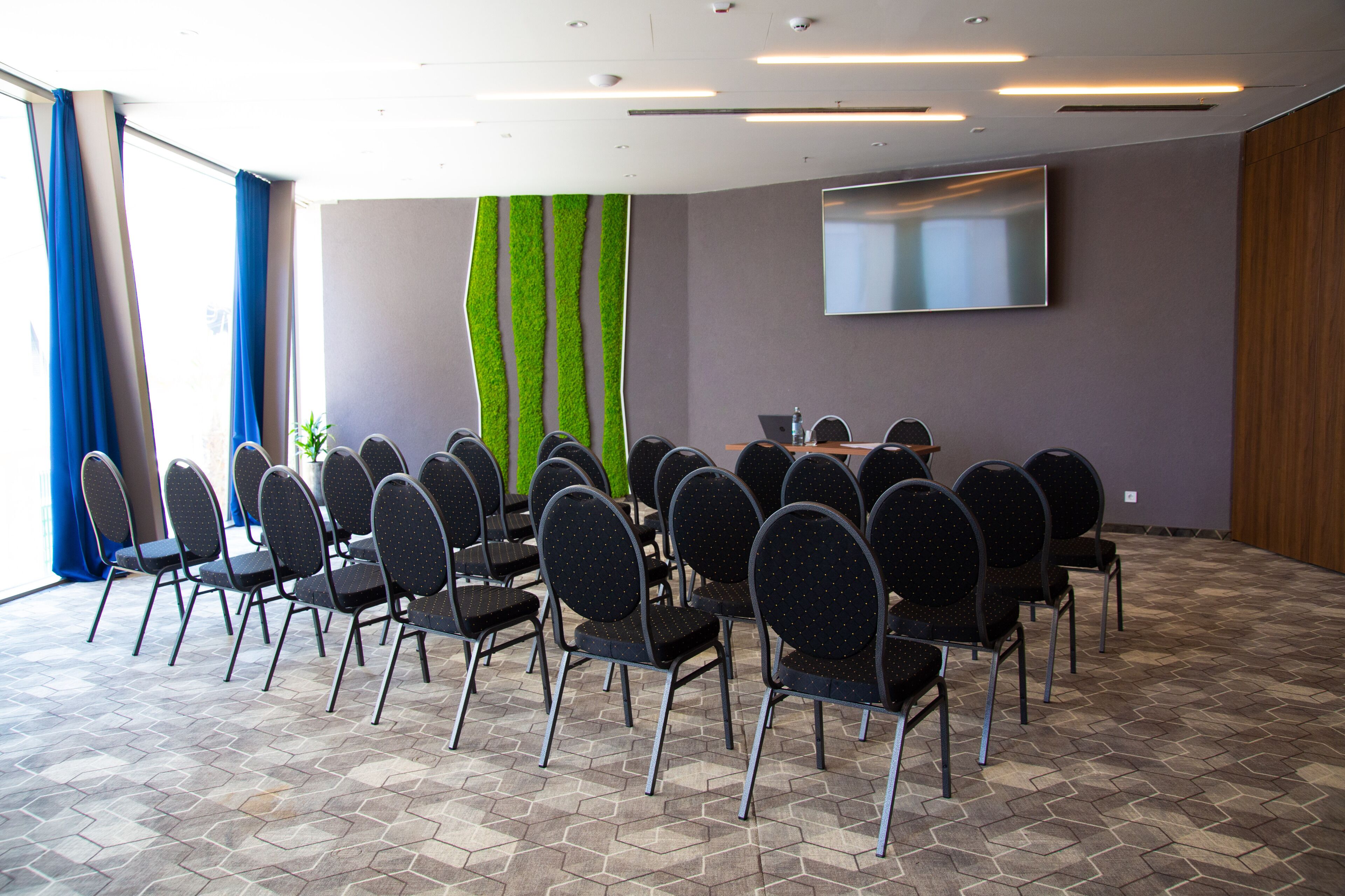 A conference room with a large table surrounded by chairs, a flat-screen TV on the wall, and a patterned floor.