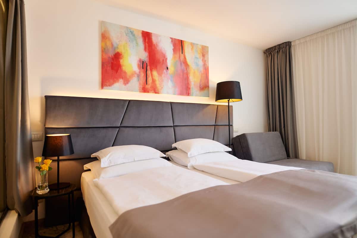 A hotel room with a large bed, a bedside table with a lamp, a wall-mounted abstract painting, and a view of the outside through curtains.