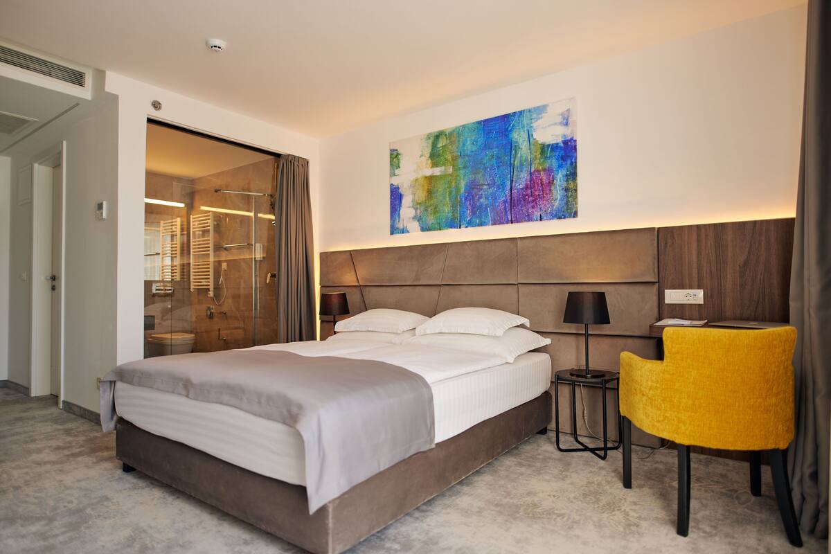 A modern hotel room with a large bed, a small desk, a chair, and a colorful abstract painting on the wall.