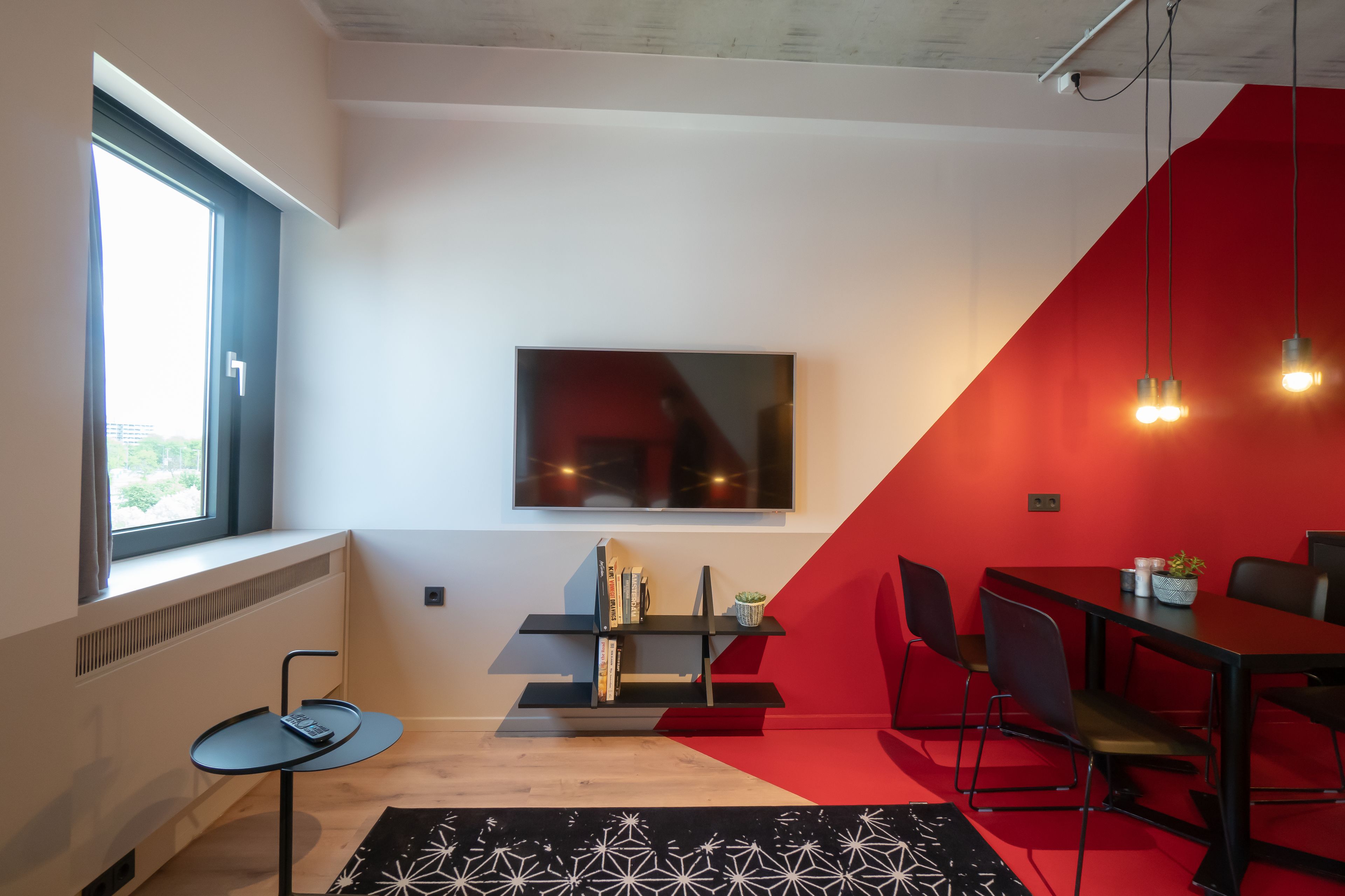 A modern room with a large window, a black and white patterned rug, a red accent wall, a black TV, and a dining area with chairs and a table.