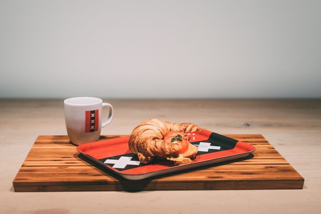 Un croissant sobre una bandeja roja con un diseño de cruz blanca, junto a una taza blanca con una franja roja y negra.