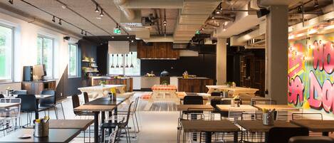 A modern restaurant with a mix of dining and kitchen areas, featuring exposed ceilings, industrial lighting, and a vibrant graffiti wall.