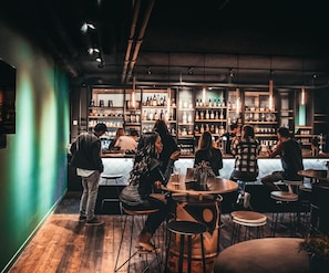 A bar with round tables and stools, shelves with bottles, and a green accent wall. - Via Suites (Diemen)