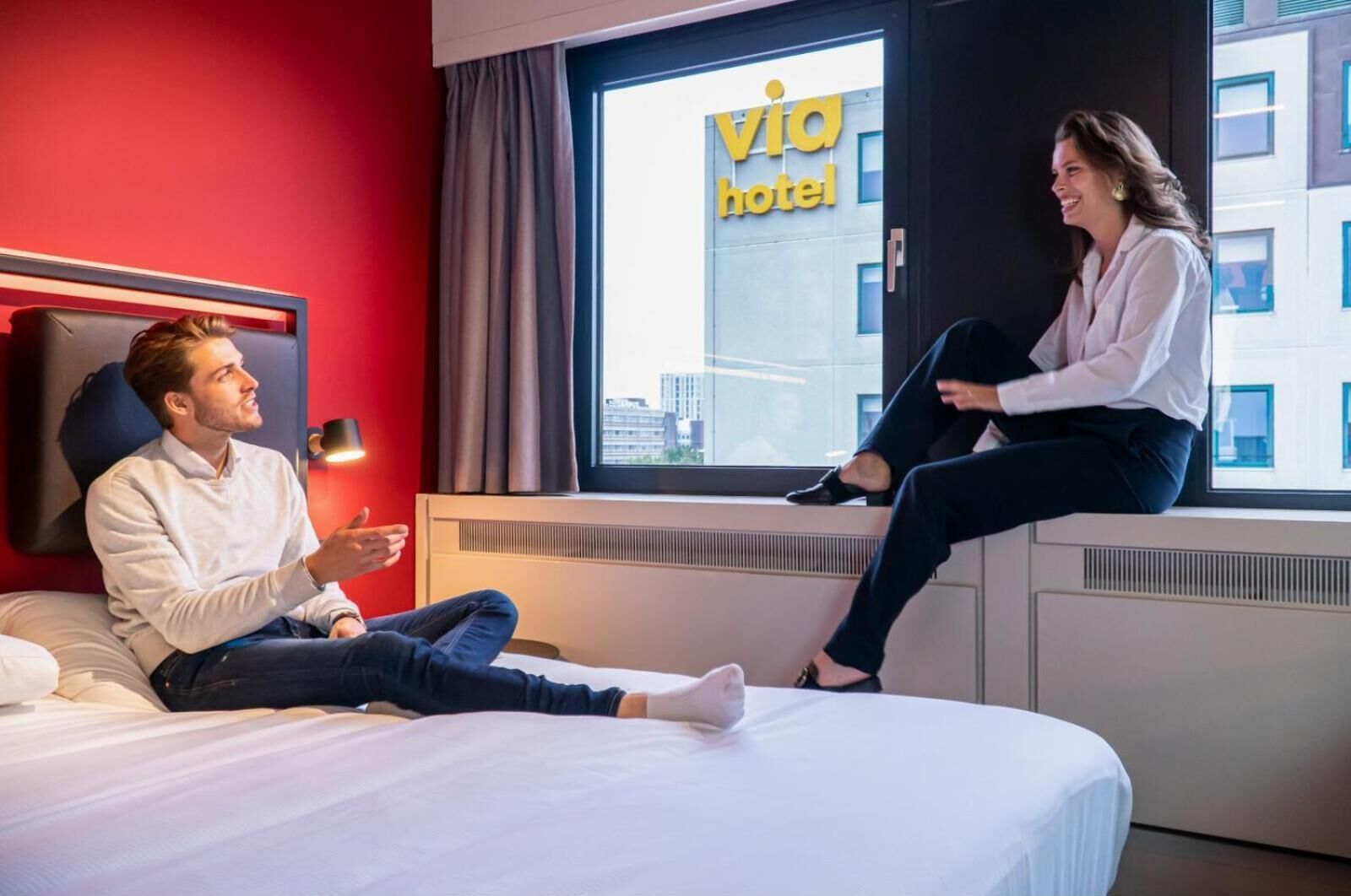 Quarto de hotel com cama, janela e vista da cidade, e um letreiro com a inscrição "via hotel".