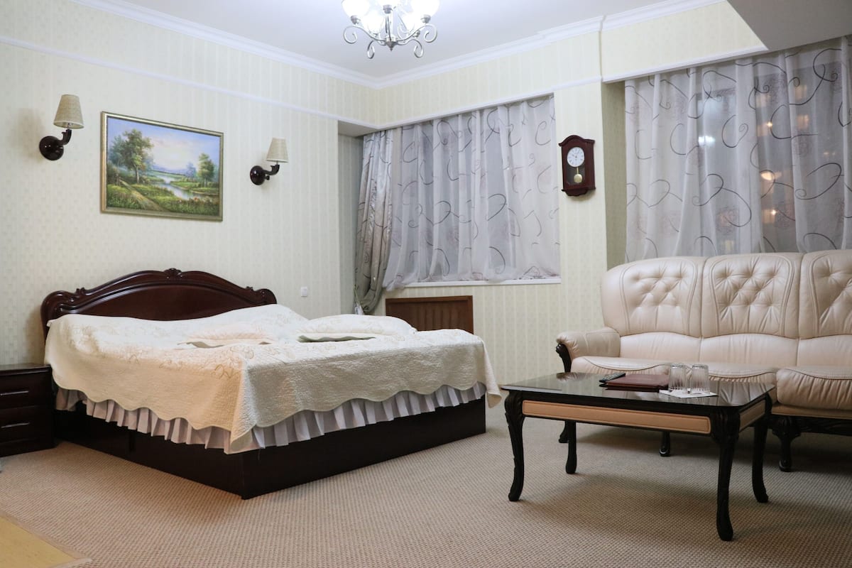 A bedroom with a bed, a sofa, a table, a painting, and a clock on the wall.