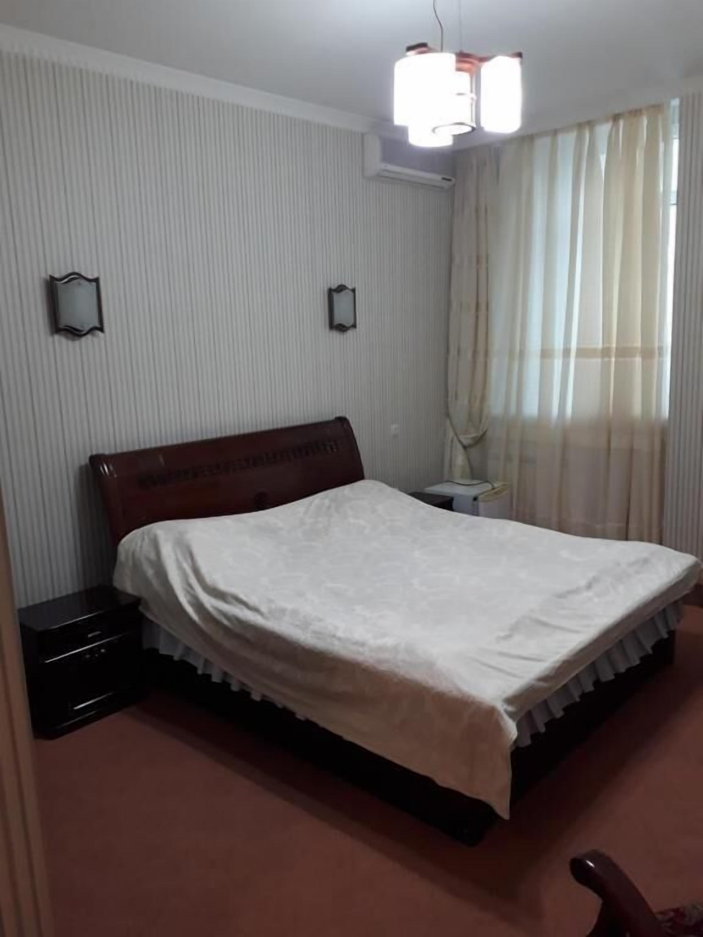 A bedroom with a single bed, a bedside table, a nightstand, a ceiling light fixture, and a window with curtains.