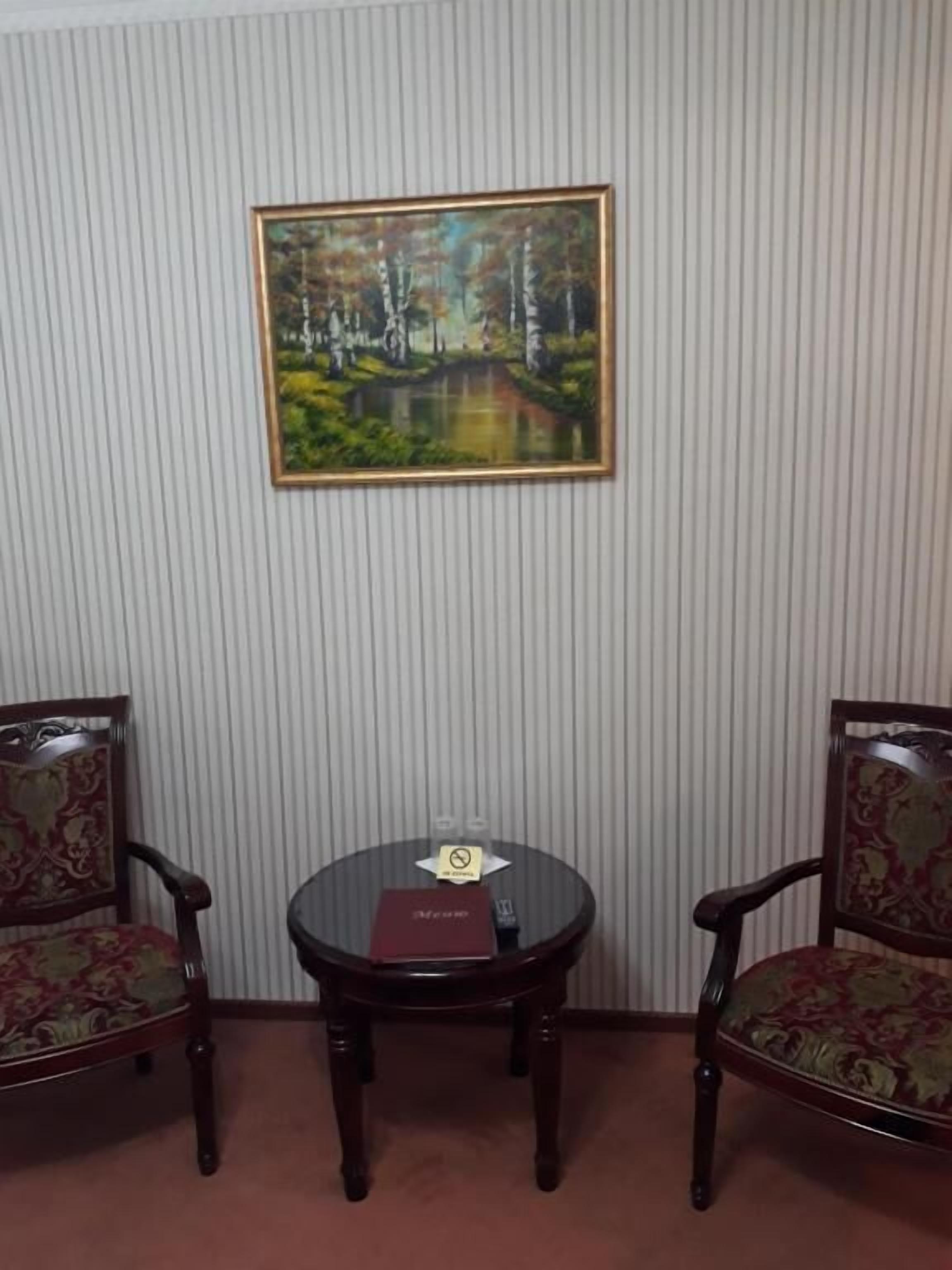A room with two wooden chairs with floral upholstery, a small round table with a red book, and a framed painting of a forest scene on the wall.