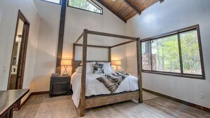 A bedroom with a four-poster bed, a nightstand, a lamp, and large windows.