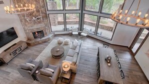 A spacious living room with a stone fireplace, large windows, and a modern chandelier.