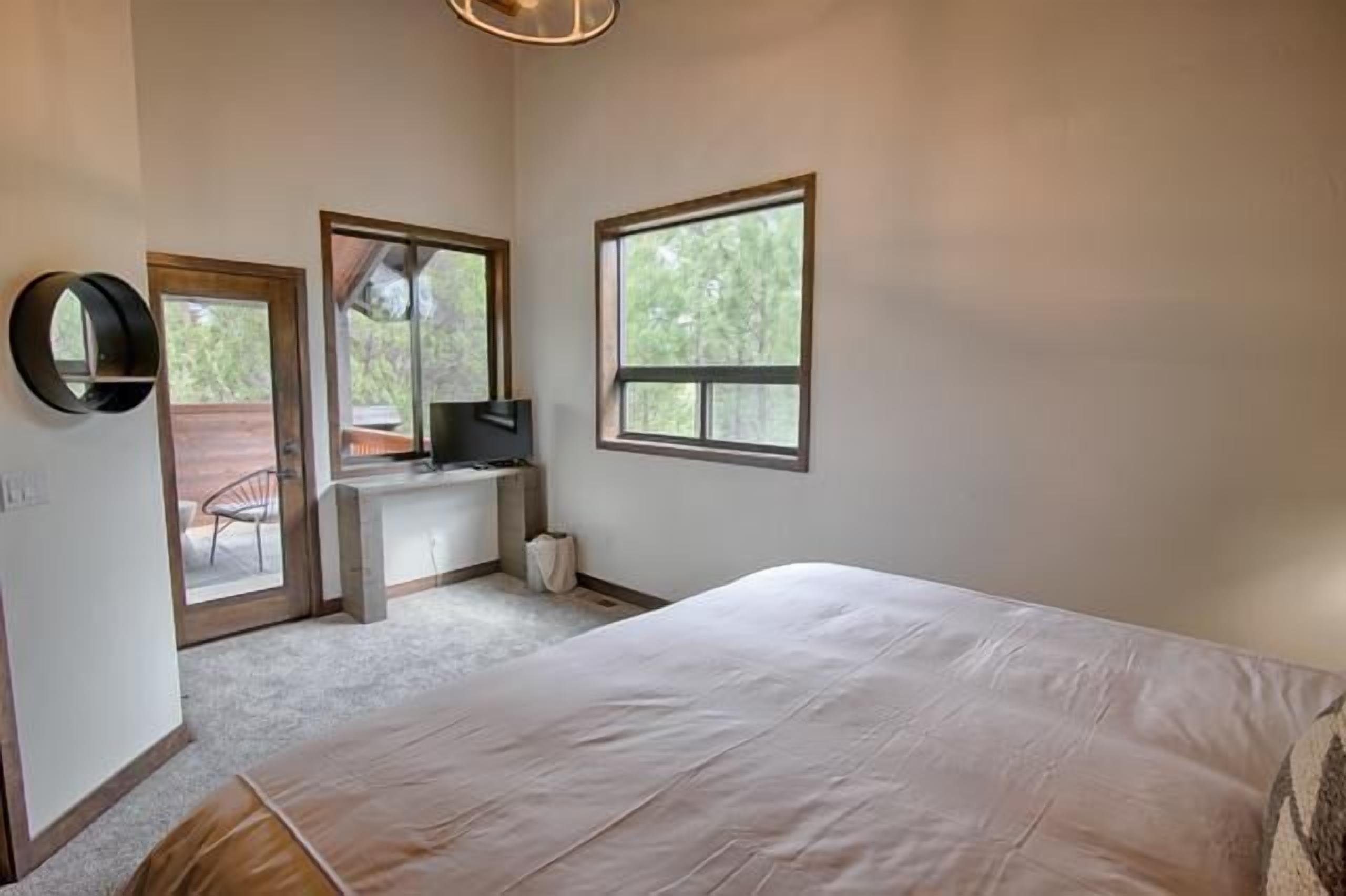 Quarto com uma cama grande, uma escrivaninha com computador, uma cadeira e duas janelas.
