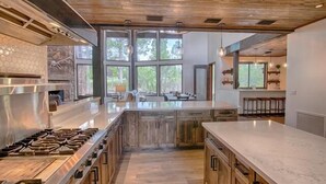 A modern kitchen with a large island, wooden cabinets, and a view of the outdoors through large windows.