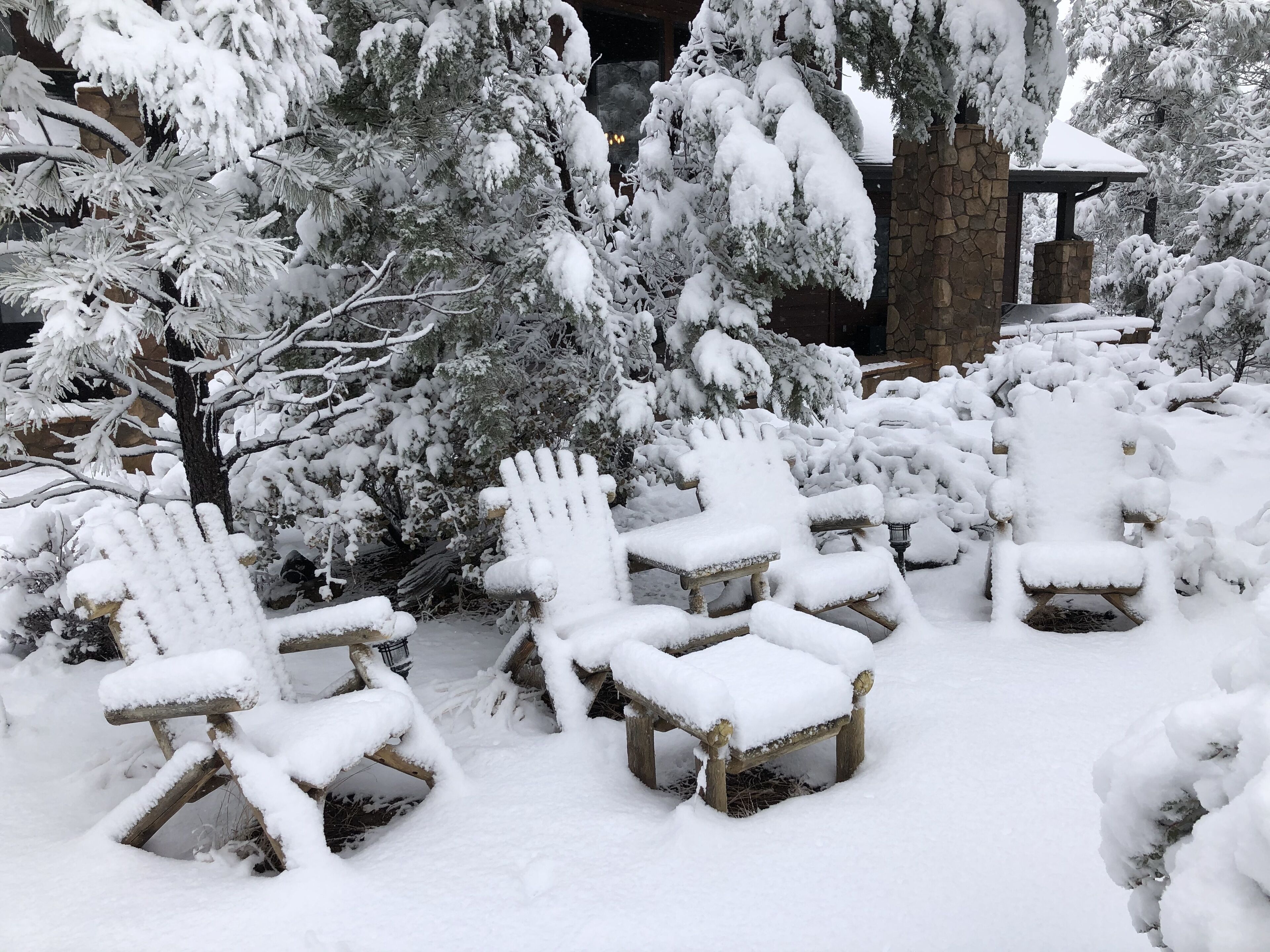 Outdoor wooden chairs covered in snow, surrounded by snow-covered trees and a stone structure.