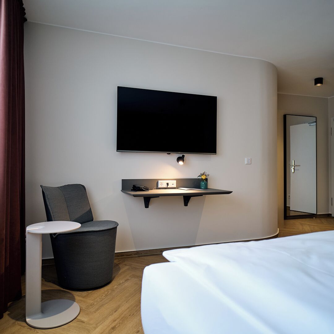 Une chambre d’hôtel avec un lit, un téléviseur fixé au mur, une petite table avec une chaise et une poubelle.