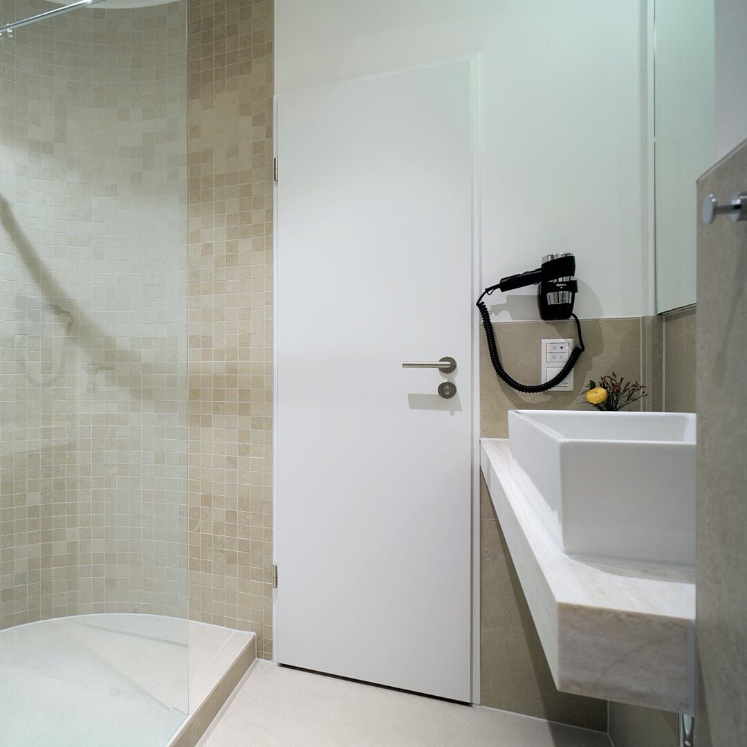 A bathroom with a shower, a white door, a hairdryer on the wall, and a sink with a marble countertop.