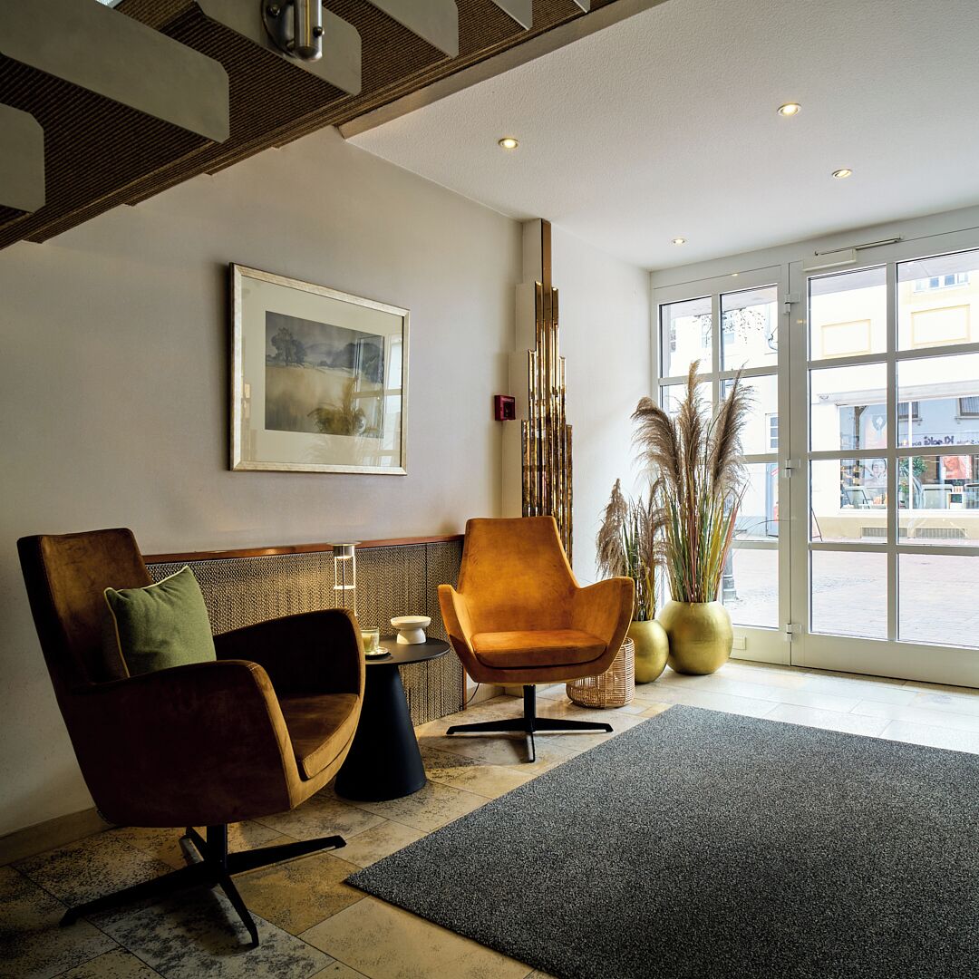 A modern lobby with a wooden bench, a single armchair, a framed artwork on the wall, and a large window with a view of the street.