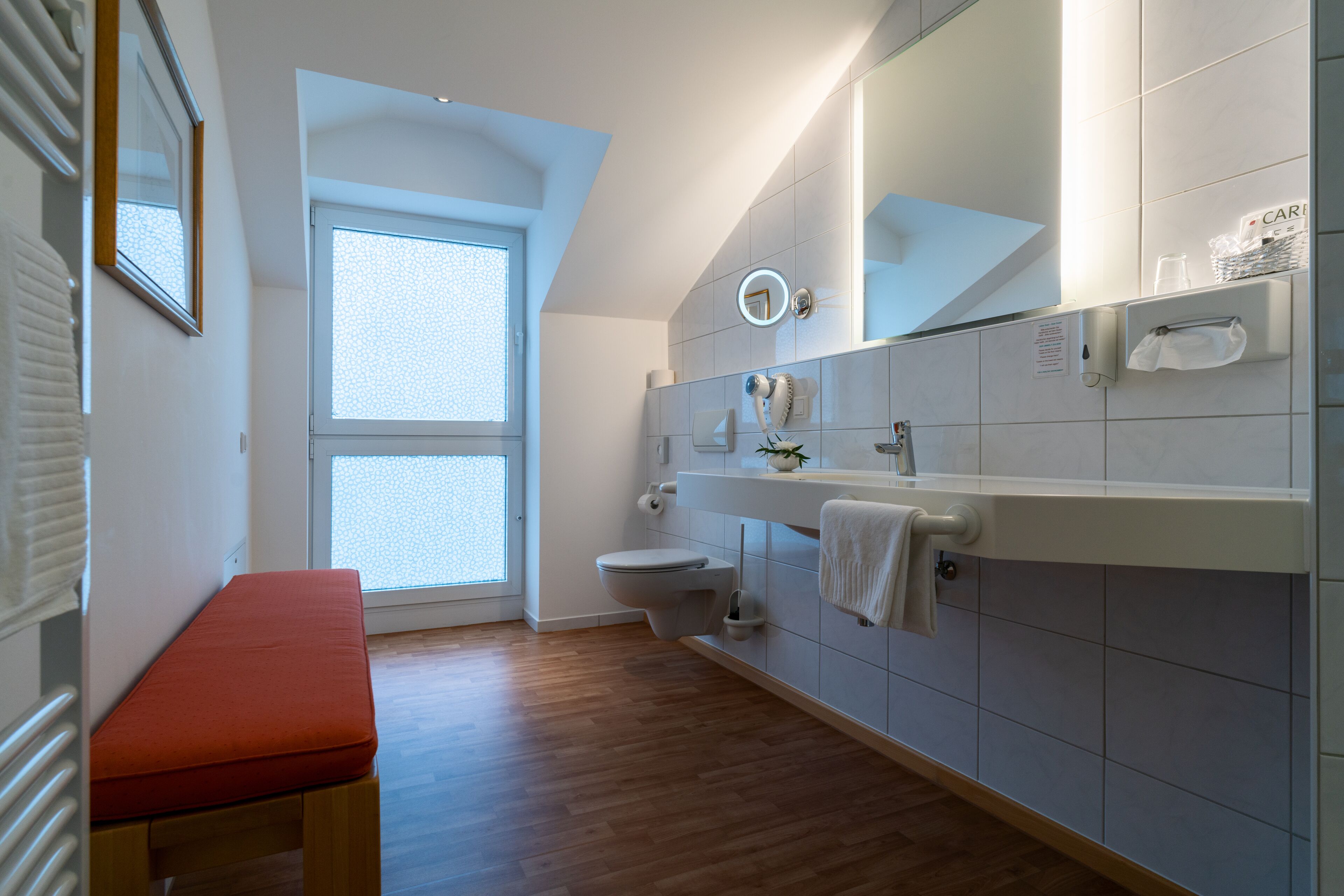 Un baño moderno con un banco de madera, un inodoro, un lavabo con un perchero para toallas y una ventana.
