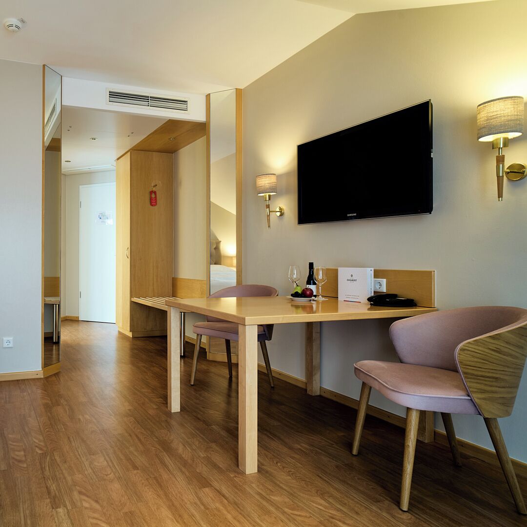 Quarto de hotel com mesa de jantar de madeira, televisão de tela plana na parede, pequena escrivaninha com luminária, uma cadeira e uma cama ao fundo.