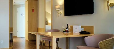 Quarto de hotel com mesa de jantar de madeira, televisão de tela plana na parede, pequena escrivaninha com luminária, uma cadeira e uma cama ao fundo.