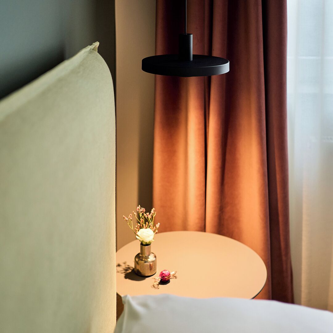 Un letto rifatto con cura, un cuscino bianco e un piccolo comodino con un vaso di fiori.