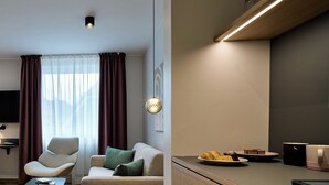 A modern living room with a sofa, armchair, and a small table. There is a kitchen area with a countertop and a dining table. - Hotel Engbert (Oelde)