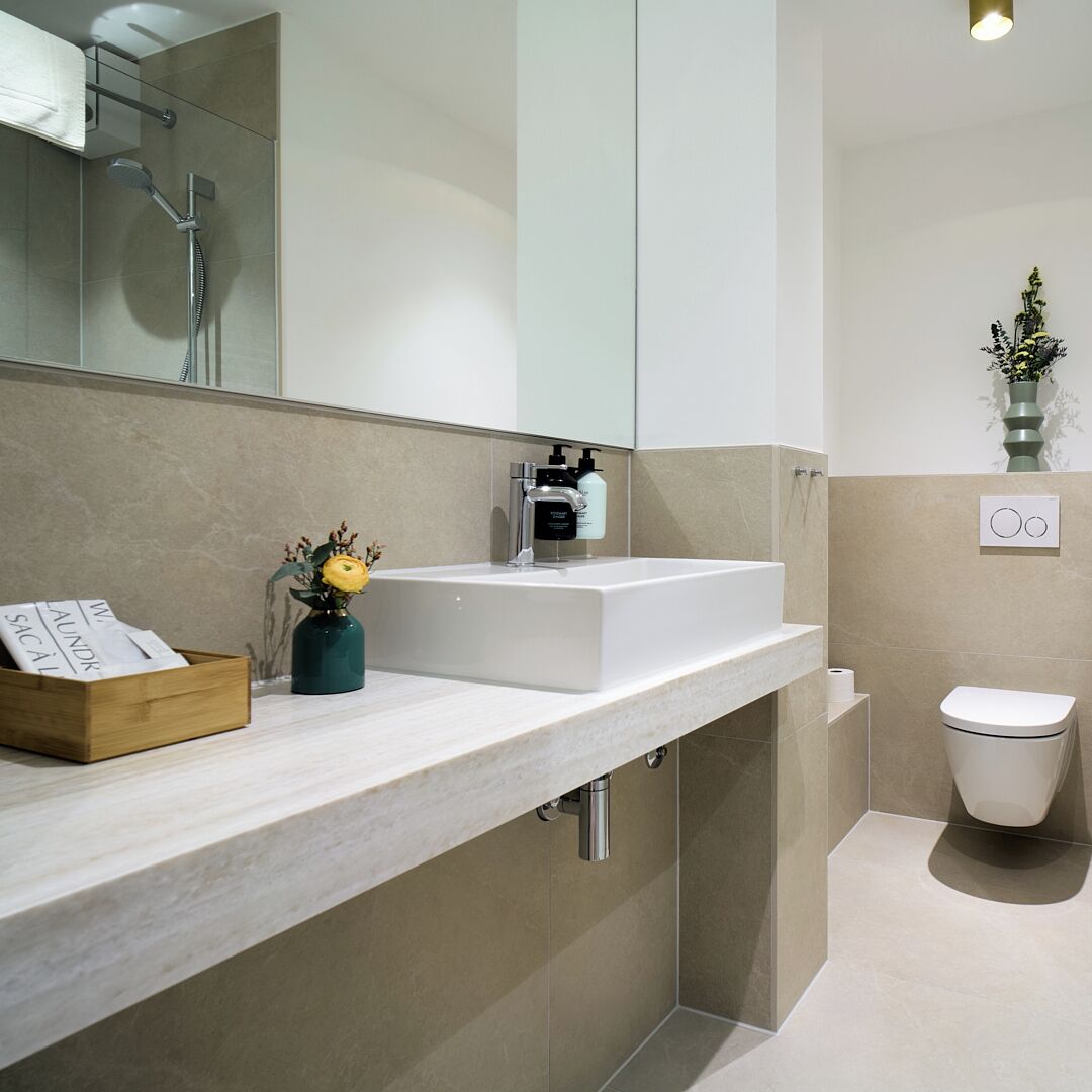 A modern bathroom with a white sink, a shower, and a toilet. There is a wooden box on the counter and a vase with flowers.