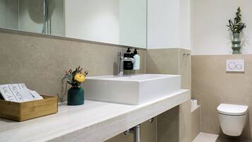 Ein modernes Badezimmer mit einem weißen Waschbecken, einer Dusche und einem WC. Auf der Ablagefläche steht eine Holzschatulle und eine Vase mit Blumen.