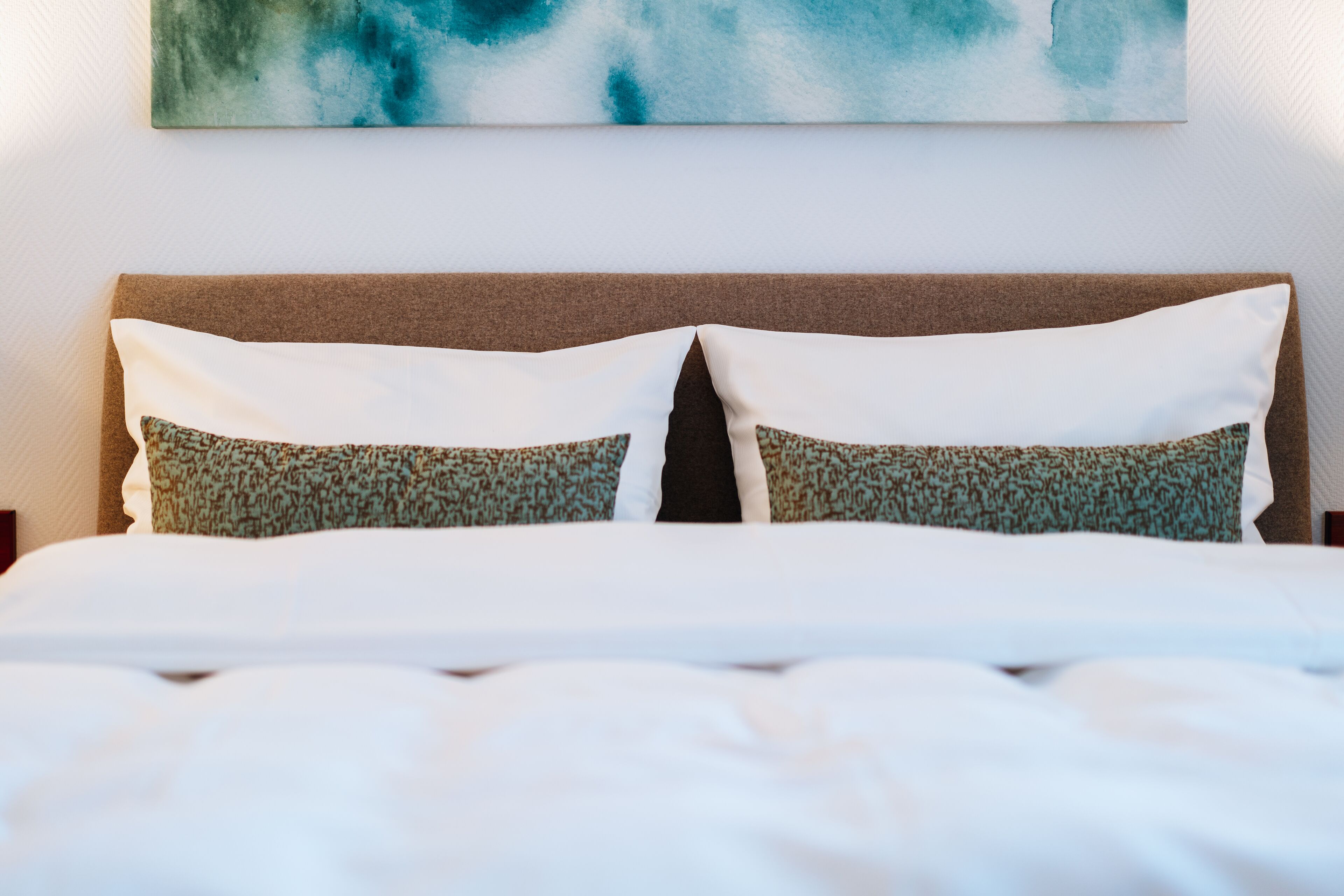 A neatly made bed with white linens and patterned pillows, a brown headboard, and a teal abstract painting on the wall.