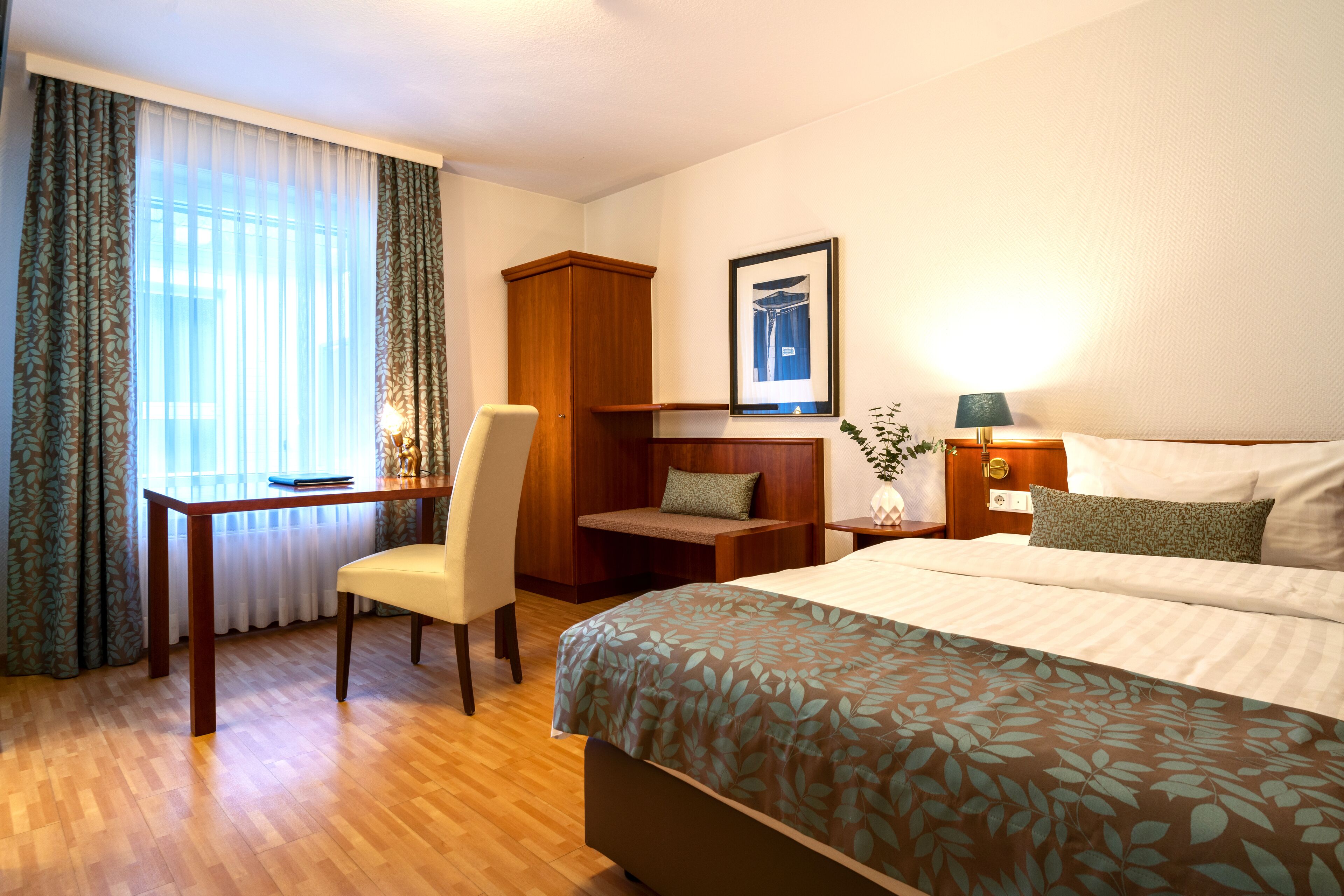 Een hotelkamer met een bed, bureau, stoel, garderobe en nachtkastje met lamp.