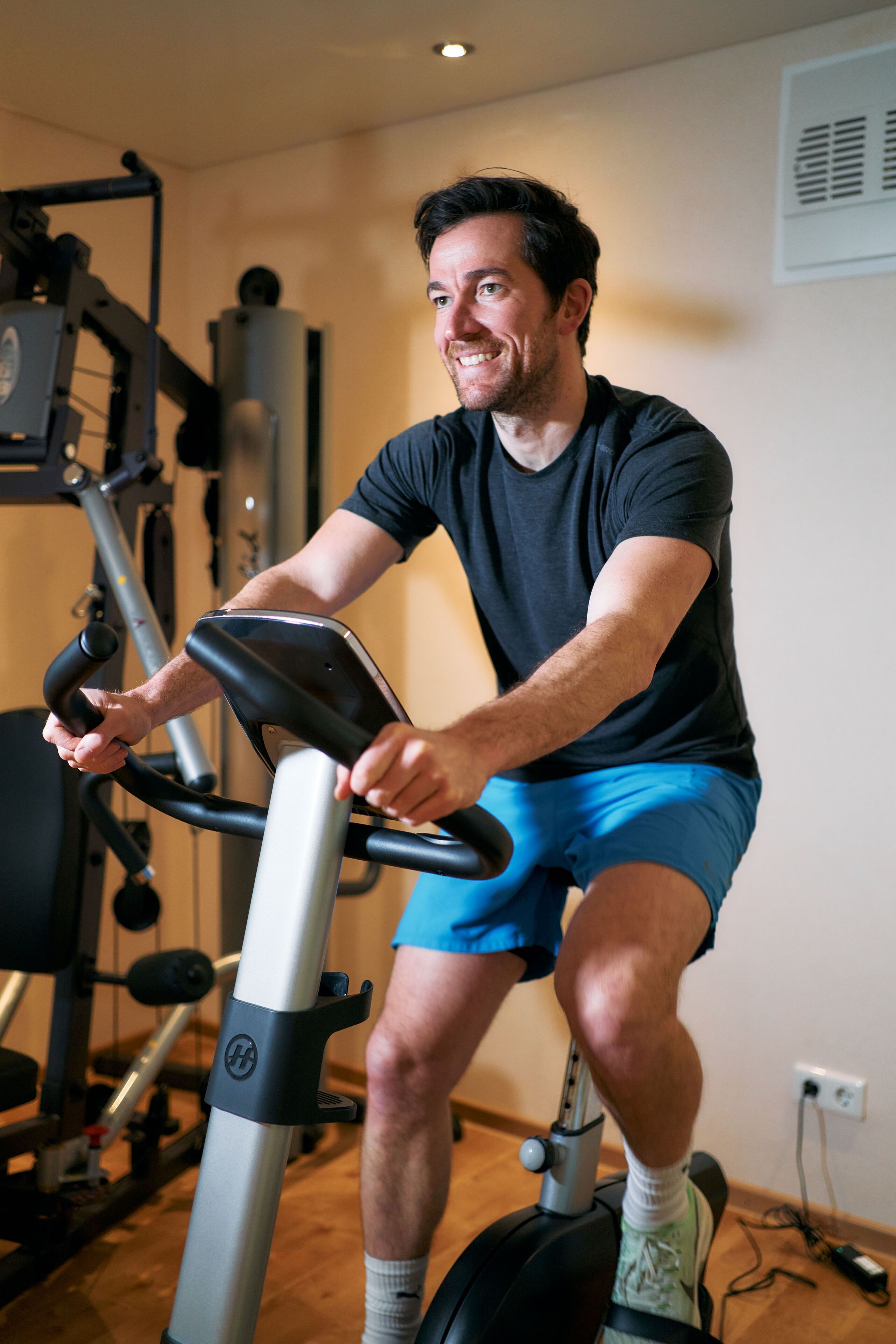 A person using an exercise bike in a room with a visible wall outlet and exercise equipment in the background.