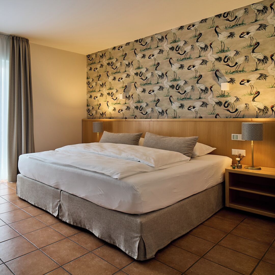 A hotel room with a large bed, two bedside tables with lamps, a wall with a bird pattern, and a window with curtains.