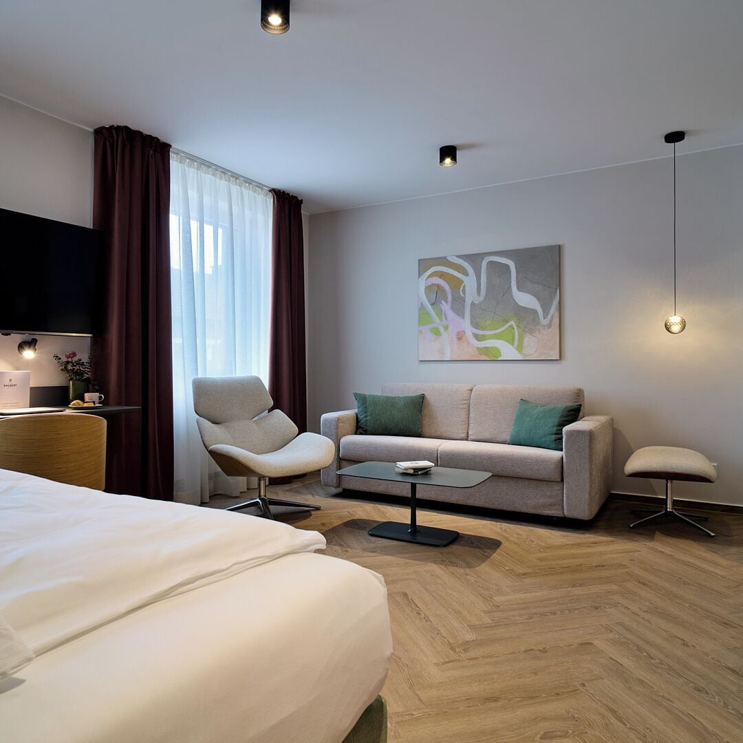 Ein modernes Hotelzimmer mit Bett, Sofa, Sessel, Couchtisch und Fernseher.
