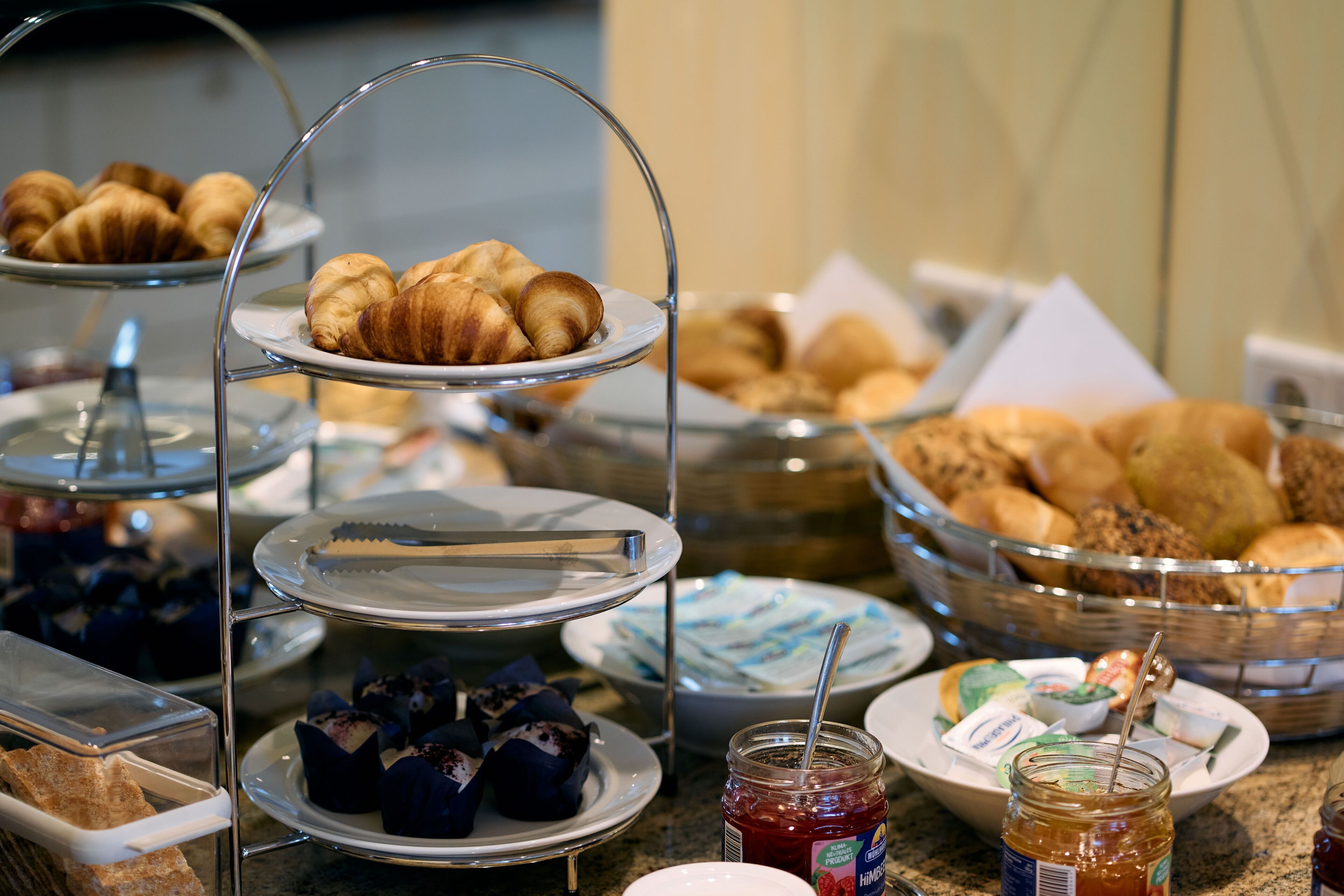A buffet with various pastries and desserts, including croissants and muffins, arranged on multiple serving stands.