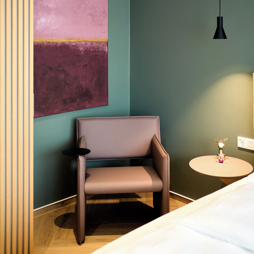 A modern room with a brown chair, a small round table with a vase, and a large abstract painting on the wall.