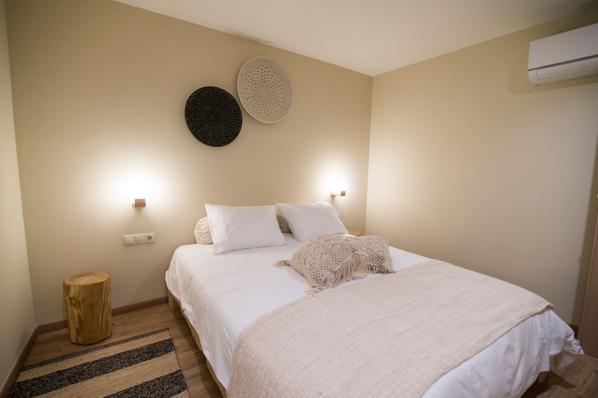 A bedroom with a bed, two wall-mounted lights, a round wall decoration, a small wooden stool, and a striped rug.