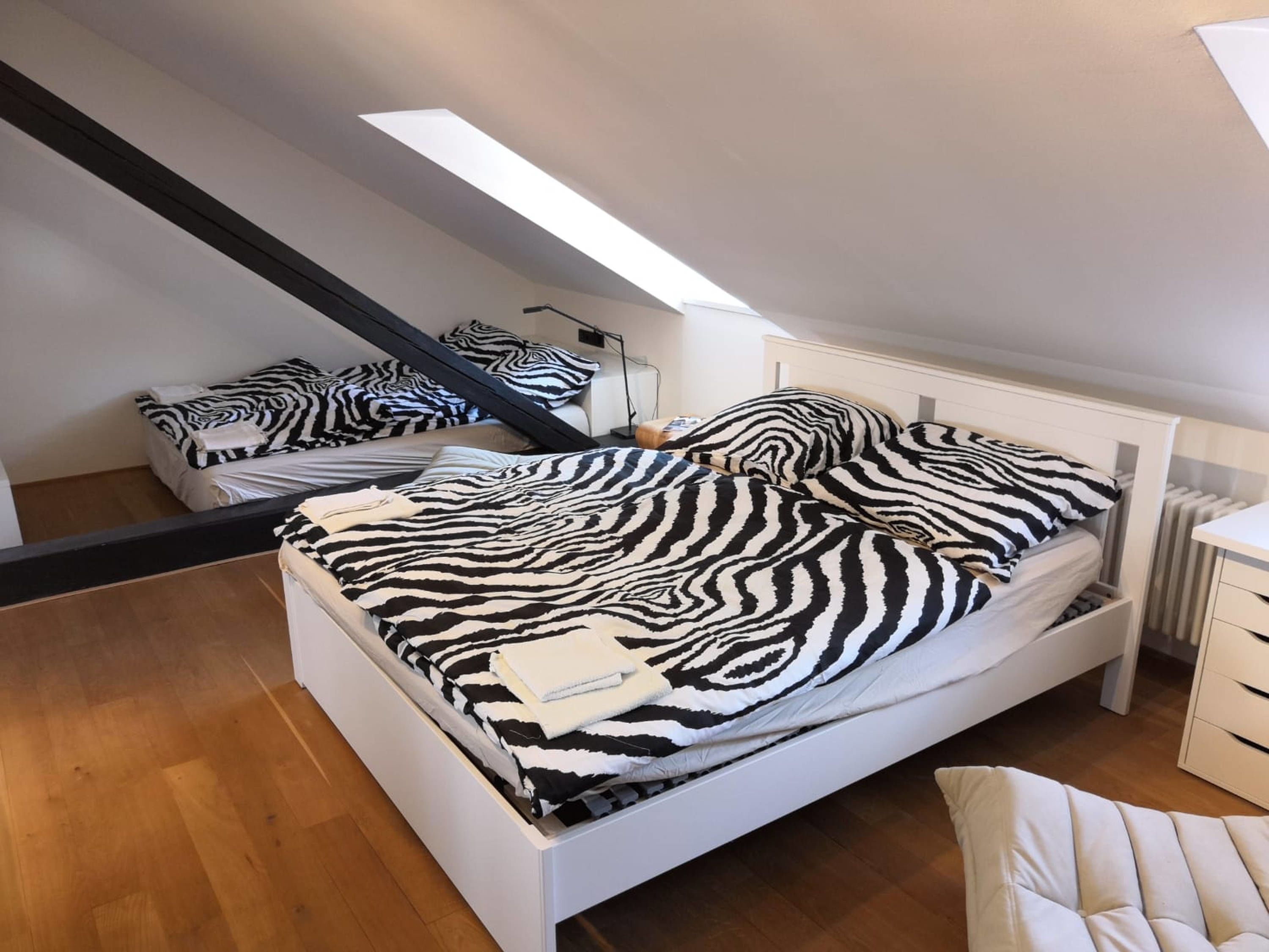 Une chambre à coucher avec un lit blanc, une literie à motifs zébrés, une commode blanche et une lucarne.